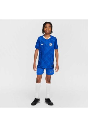 Barn iført blå Chelsea-fodboldtrøje med hvide sokker og sorte fodboldstøvler, der står mod en ensartet lys baggrund.