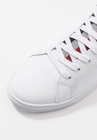 Baskets blanches en cuir avec les lacets blancs sur un tissu noir, blanc et rouge, couture détaillée et semelle en caoutchouc texturé sur fond blanc.