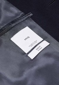 Vnitřní podšívka černého saka s dvěma bílými štítky s uvedenou značkou MNG, velikostí střední, slim fit a materiálem z vlněné směsi.