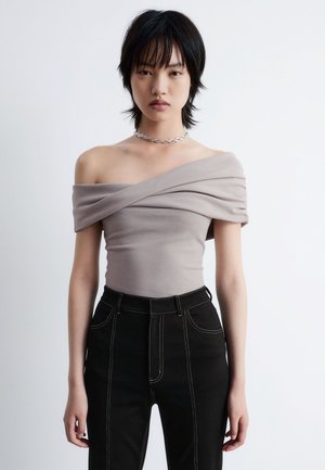 Beiger Off-Shoulder-Top mit gerafftem Stoffdetail, kombiniert mit hochgeschnittenen schwarzen Jeans mit Kontrastnähten und einer eleganten Silhouette.