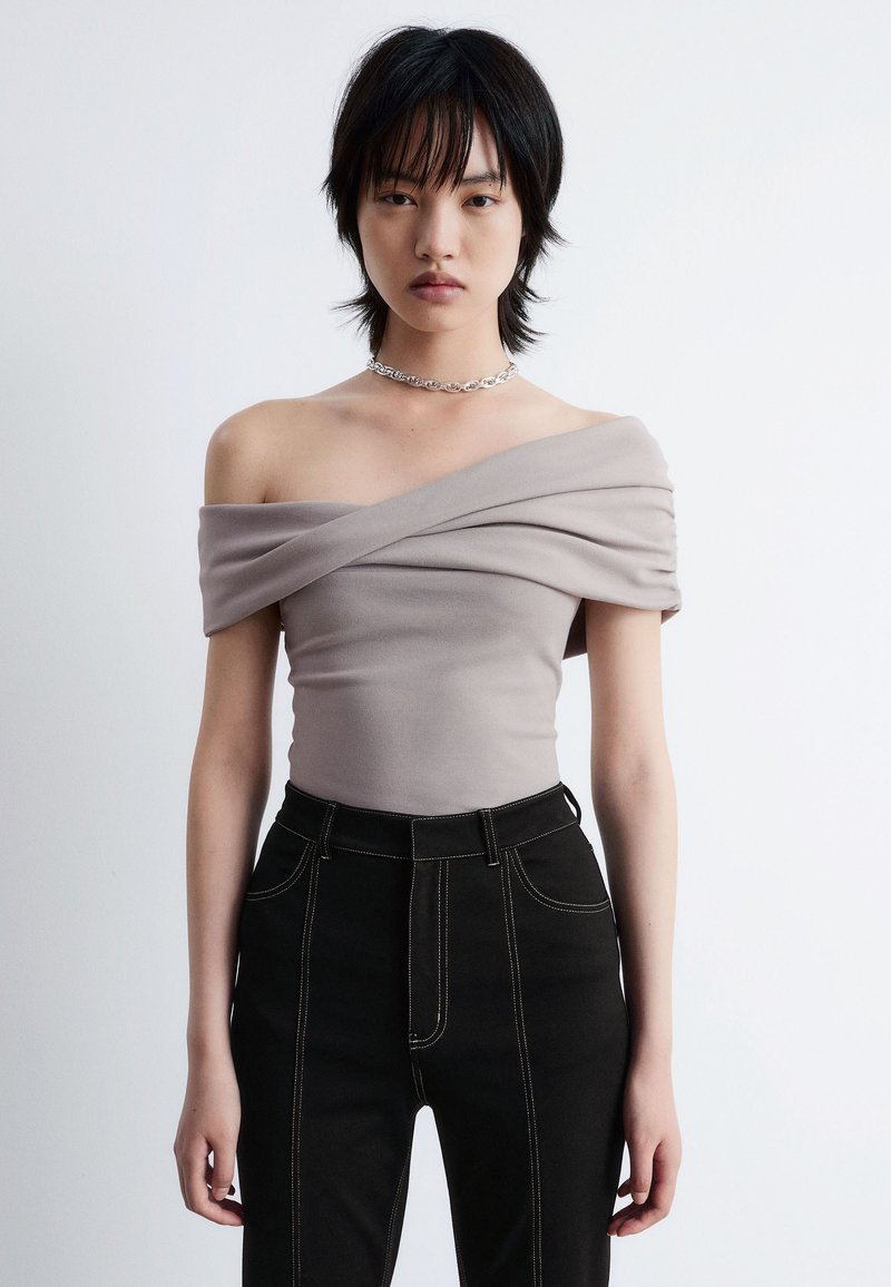 Beiger Off-Shoulder-Top mit gerafftem Stoffdetail, kombiniert mit hochgeschnittenen schwarzen Jeans mit Kontrastnähten und einer eleganten Silhouette.