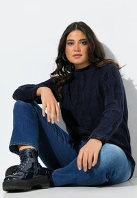 Marineblauwe gebreide trui met getextureerde golfpatronen, hoge hals en lange mouwen, gecombineerd met donkerblauwe jeans en glanzende zwarte militaire laarzen.