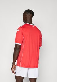 Punane spordijersey lühikeste varrukatega, valgete detailidega ja tekstuuriga mustriga külgedel. Mudel kannab valgeid lühikesi pükse.