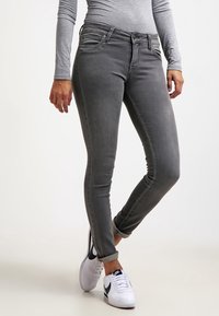 Osoba v úzkých tmavě šedých džínách s ohrnutými kotníky, šedém dlouhém rukávu a bílých teniskách Nike s černým logem swoosh.