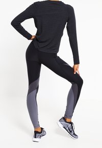 Top negro de manga larga con hombros fruncidos, combinado con leggings ajustados que presentan paneles en gris oscuro. Se lleva con zapatillas deportivas en blanco y negro.