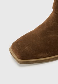 Bottine en suède marron avec un bout carré, dotée d'une semelle en caoutchouc beige et d'une texture lisse. Pas de matériel visible ni de motifs.