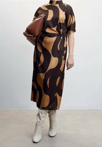 Robe à motif brun et doré avec une taille cintrée, longueur genoux, manches courtes. Associée à des bottes blanches moulantes et un sac à main marron.