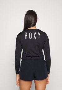 Čierna dlhá rukávová rash guard s nápisom "ROXY" v bielej farbe na zadnej strane, v kombinácii s čiernymi šortkami, vyrobená z hladkej, elastickej tkaniny.