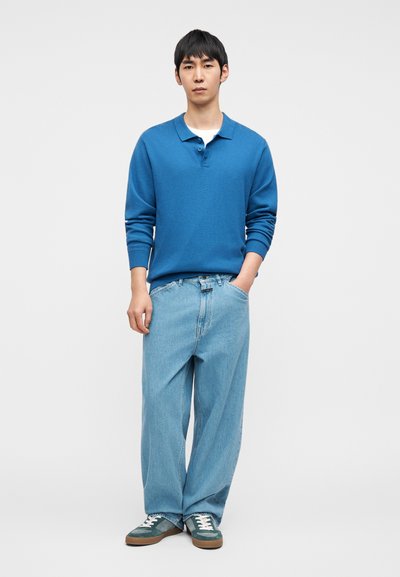 Μπλε πόλο μπλούζα με μακριά μανίκια και γιακά, σε συνδυασμό με ανοιχτόχρωμα χαλαρά τζιν και πράσινα αθλητικά παπούτσια με λευκά κορδόνια.