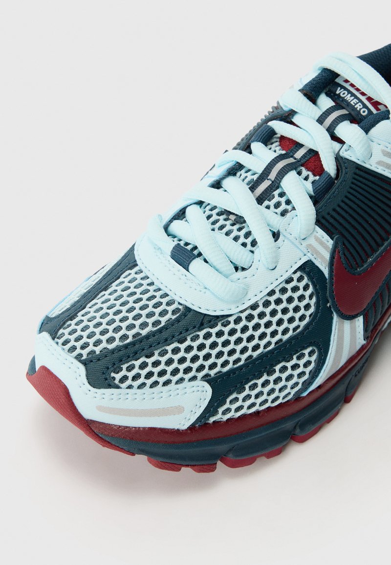 Nike hardloopschoen met een lichtblauwe mesh bovenkant, marineblauwe accenten en een rode hiel. Textuurontwerp en zichtbaar vetersysteem.