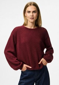 Burgundy ylipitkä neulepaita, jossa on pyöreä kaula-aukko, pitkät hihat ja ribbireunus. Valmistettu pehmeästä, teksturoidusta materiaalista. Yhdistetty tummiin farkkuihin.