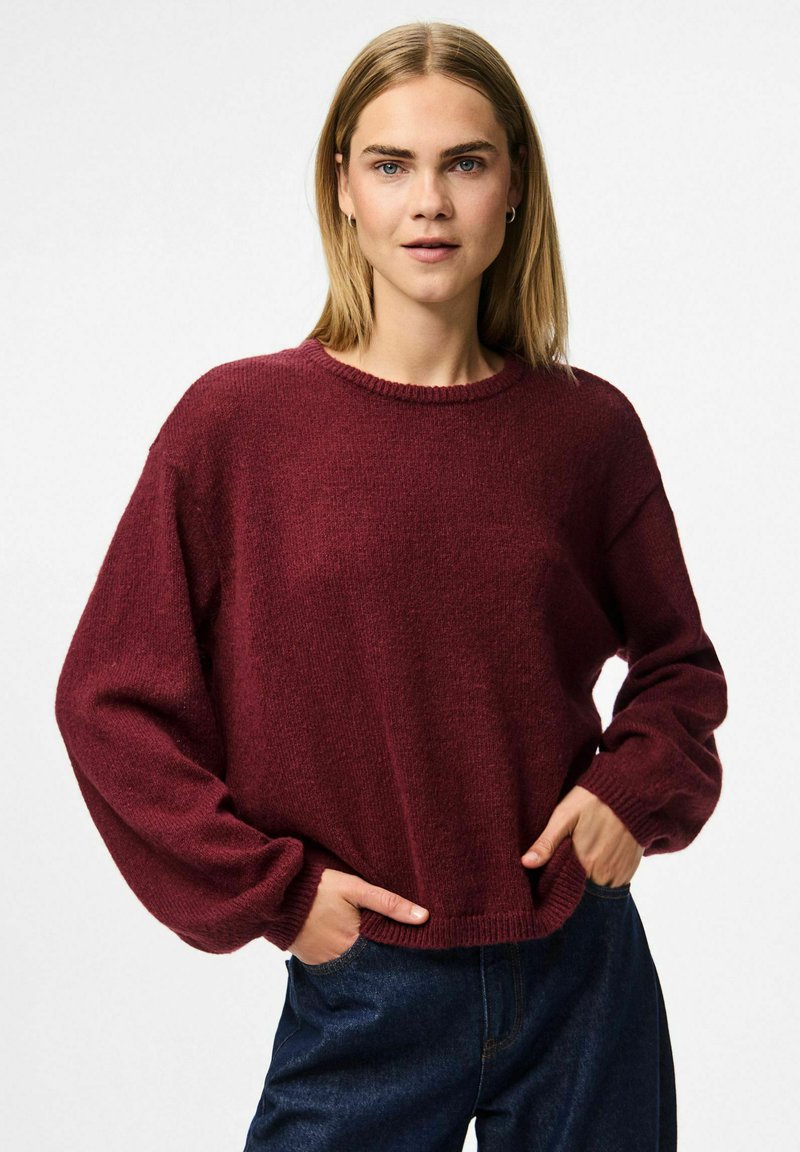 Burgundy ylipitkä neulepaita, jossa on pyöreä kaula-aukko, pitkät hihat ja ribbireunus. Valmistettu pehmeästä, teksturoidusta materiaalista. Yhdistetty tummiin farkkuihin.