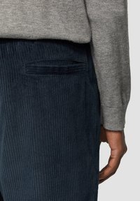 Pantalon en velours côtelé bleu marine avec des côtes verticales et une poche arrière avec un surpiqure. Porté avec un pull texturé gris clair.