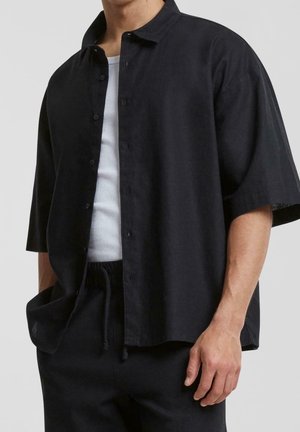 Mand iført sort kortærmet skjorte med knapper over hvid ribbet tanktop og sorte bukser med snøre, stående med den ene hånd i lommen.