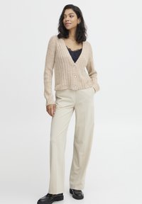Cardigan beige côtelé avec un col en V, une fermeture à boutons et des boutons en perle, porté sur un top en dentelle noir, assorti avec un pantalon crème et des chaussures noires.
