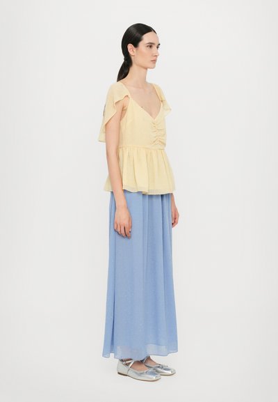Gelbes Peplum-Top mit kurzen, rüschierten Ärmeln und Vorderraffungen, kombiniert mit einem hellblauen Maxikleid und silbernen flachen Schuhen.