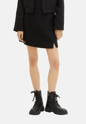 Kvinde iført en sort tekstureret mininederdel med forreste slidser, matchende cropped jakke og sorte snørestøvler med chunky såler.