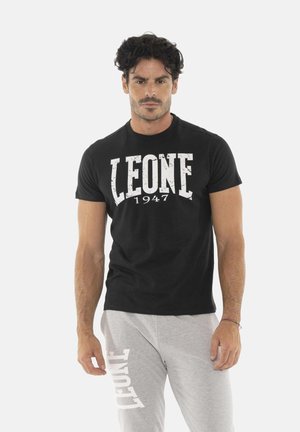 Homme aux cheveux bouclés foncés portant un t-shirt noir "LEONE 1947" et un pantalon de survêtement gris clair avec l’inscription "LEONE" sur la jambe gauche.