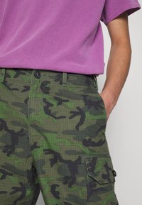 Vihreät maastokuvioiset cargo-housut, joissa on ruudullinen tekstuuri, mustat yksityiskohdat, kaksi sivutaskua ja napillinen suljenta. Yhdistetty violettiin paitaan.