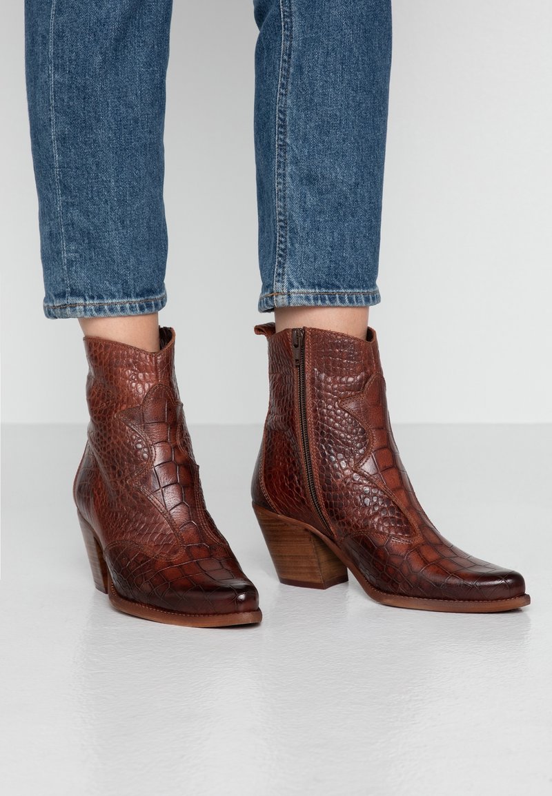 Bruna läderankelstövlar med ett texturerat krokodilmönster, spetsig tå och klack i staplad design. Sidsdragkedja. Bärs tillsammans med uppvikta blå jeans.