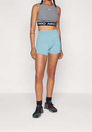 Kobieta stoi, ubrana w szary crop top Nike, jasnoniebieskie sportowe szorty z wysokim stanem, czarne skarpetki i czarne buty sportowe na jednolitym tle.
