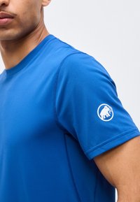 Κοντινό πλάνο ενός ατόμου που φορά μπλε πουκάμισο με κοντά μανίκια και λευκό λογότυπο μαμούθ στο αριστερό μανίκι, σε απλό φόντο.