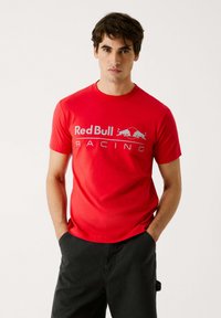 Rød kortærmet t-shirt lavet af glat stof med "Red Bull Racing" grafik og logo i hvidt på tværs af brystet.