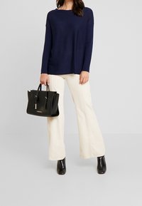 Granatowa bluzka z długim rękawem, kremowe spodnie o szerokich nogawkach, czarna skórzana torebka z złotymi akcentami, oraz czarne botki na obcasie. Gładkie tekstury, casualowy styl.