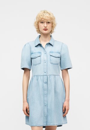 Junge Person mit kurzen lockigen blonden Haaren, die ein hellblaues Jeanskleid mit kurzen Ärmeln und Brusttaschen trägt.