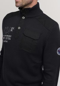 Črn pleteni pulover z visokim ovratnikom, ki ima gumbno zapenjanje, ramenske aplikacije in žep na prsih. Izvezen logotip.