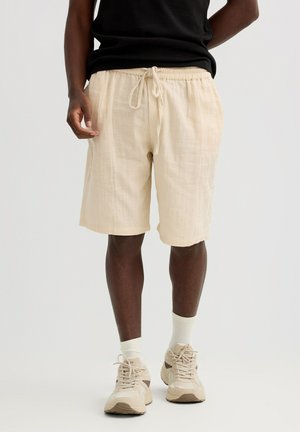 Hombre vistiendo pantalones cortos beige con cordón, calcetines blancos, zapatillas beige y una camiseta negra sobre un fondo liso claro.