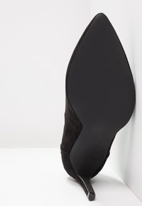 Chaussure à talon haut pour femme en daim noir inclinée vers le haut, montrant un bout pointu et la semelle sur fond blanc.