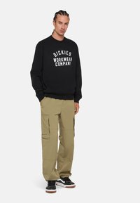 Black sweatshirt with white "Dickies Workwear Company" graphic, paired with khaki cargo pants featuring side pockets and a relaxed fit. Black sneakers.