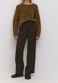 Kobieta w brązowym, puszystym swetrze, ciemnych brązowych szerokich spodniach i czarnych butach wsuwanych z futrzanym podbiciem stoi przy białej ścianie.