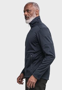 Navy softshell jacket with high collar, zippered pockets, and a fitted design. Features smooth texture and subtle branding on the arm.