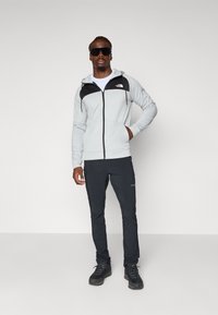 Light grey zip-up hoodie with black accents, paired with dark pants and black hiking boots. Features a front logo and a hood.