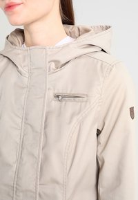 Beżowa kurtka z kapturem, z kieszenią na piersi na zamek, miękką teksturą i detalami szwów, z małą łatką z logo na rękawie.