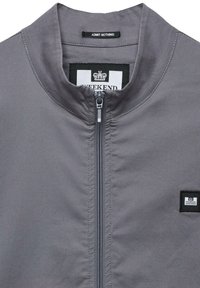 Grå jakke med høj krave, frontlynlås og lille mærkebadge. Stoffet ser glat ud med minimal tekstur.