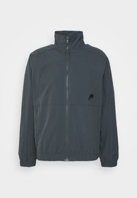 Donkergrijze ritsjack met een hoge kraag, elastische manchetten en een klein zwart logo op de borst. Glad materiaal en eenvoudig ontwerp.