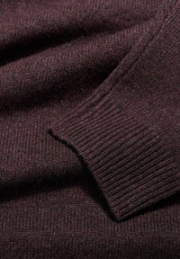 Burgundijas adīta džempera detaļa ar ribotu piedurkni un maigu tekstūru, izceļot dziļu toņu sajaukumu ar minimālu redzamu rakstu.