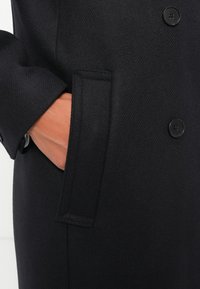 Cappotto di lana nero con una texture liscia, una tasca laterale e bottoni neri rotondi lungo la parte anteriore. Primo piano del tessuto e del dettaglio della tasca.