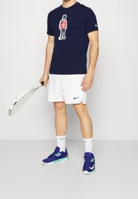 Námořnické tričko s grafickým potiskem, bílé šortky a modré sportovní boty. Drží tenisovou raketu, vystaveno na jednobarevném pozadí.