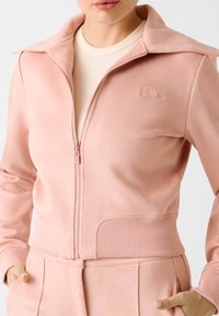 Veste zippée rose avec col, poignets et taille côtelés. Logo brodé sur la poitrine. Tissu au toucher lisse et doux.