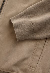 Beige Jacke mit weichem, wildlederähnlichem Material, gerippten Bündchen, einem glatten Reißverschluss und dezenten Nahtdetails auf dem Stoff.