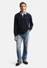 Marineblauw polo met lange mouwen en een lichtblauwe kraag, gecombineerd met vervaagde blauwe spijkerbroek en bruine sneakers. Voorzien van een geborduurd logo op de borst.