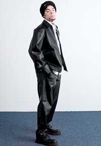 Veste en cuir noir et pantalon à la texture lisse, associés à une chemise blanche et un bonnet en tricot noir. Des chaussures noires épaisses complètent le look.