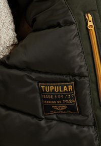 Olivově zelená prošívaná bunda s ribrovou texturou, která má zlatý zip a černou nálepku s nápisem "TUPULAR" a dalšími detaily.