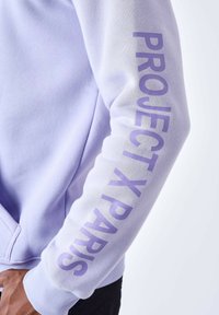 Lavendel sweatshirt med lange ærmer; har fed "PROJECT X PARIS" tekst langs venstre ærme i en matchende farve. Blød tekstur.