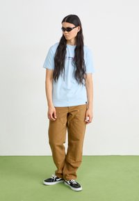 Light blue short-sleeve t-shirt with logo, paired with brown wide-leg pants and black sneakers. Model wears black sunglasses. Green floor.