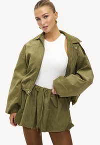 Femme portant une veste ample vert olive et une jupe taille haute assortie, avec un haut blanc sans manches et des boucles d'oreilles créoles dorées.